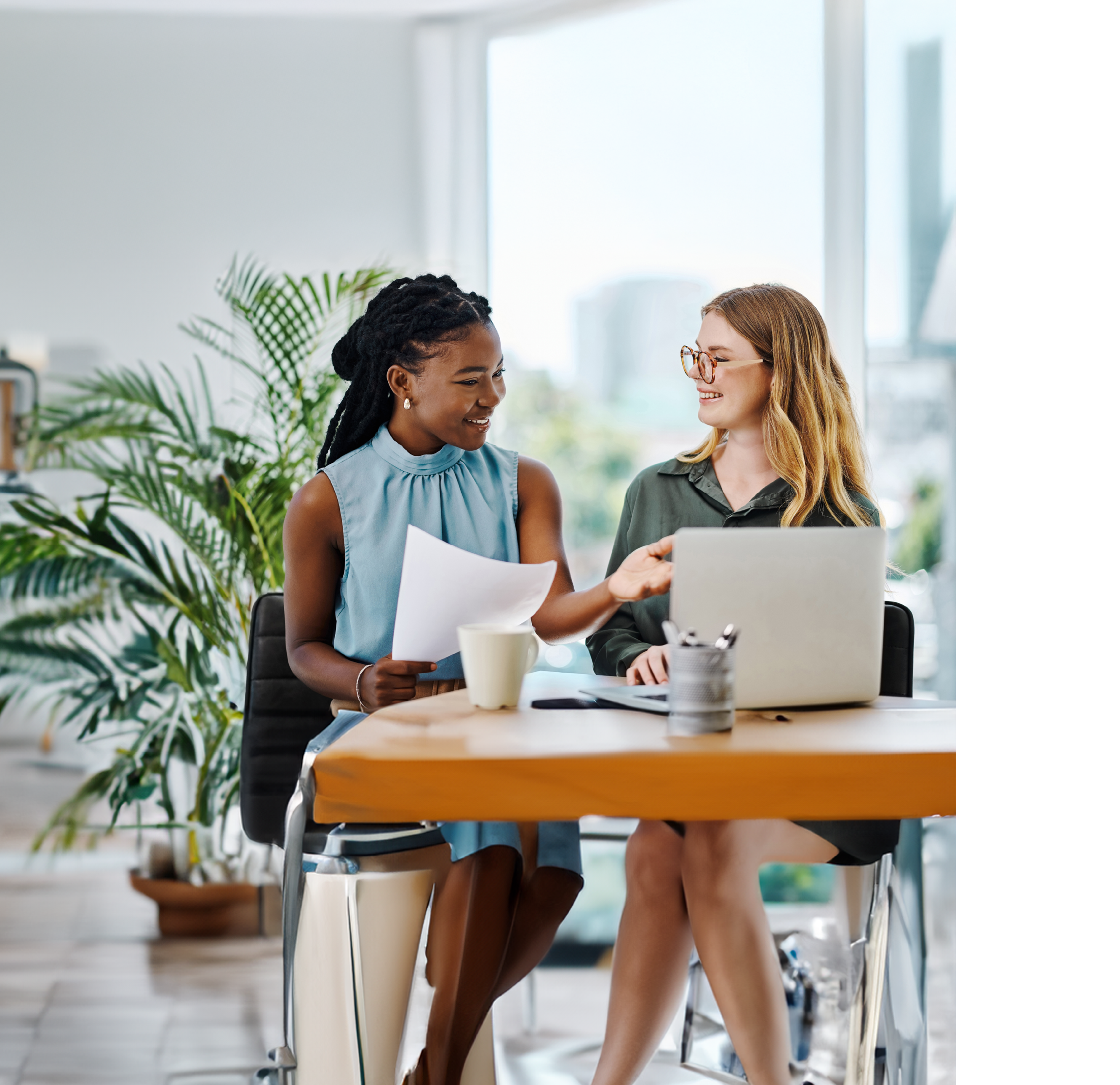 Two smiling businesswomen going through paperwork while using a laptop together in an office