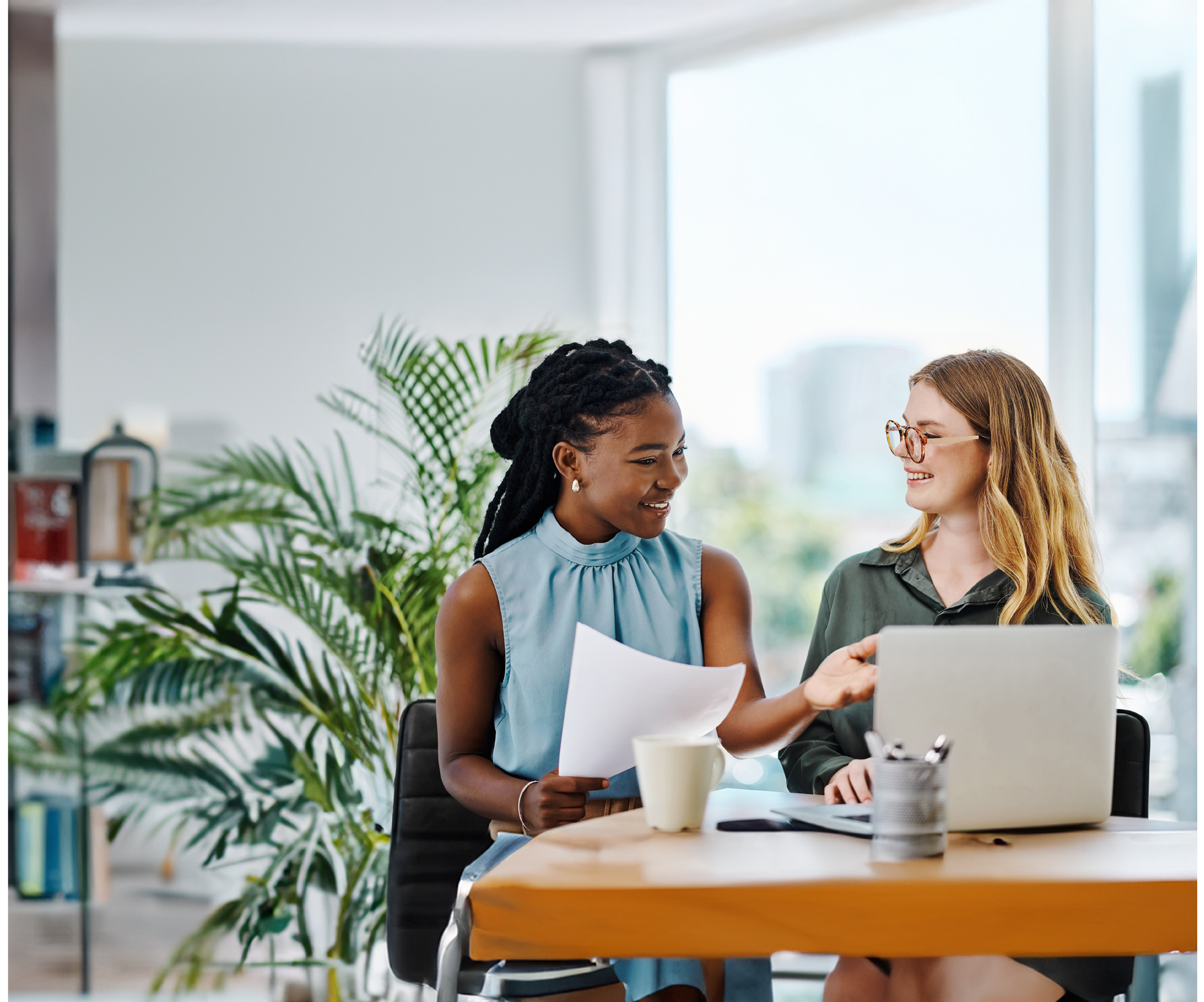 Two smiling businesswomen going through paperwork while using a laptop together in an office