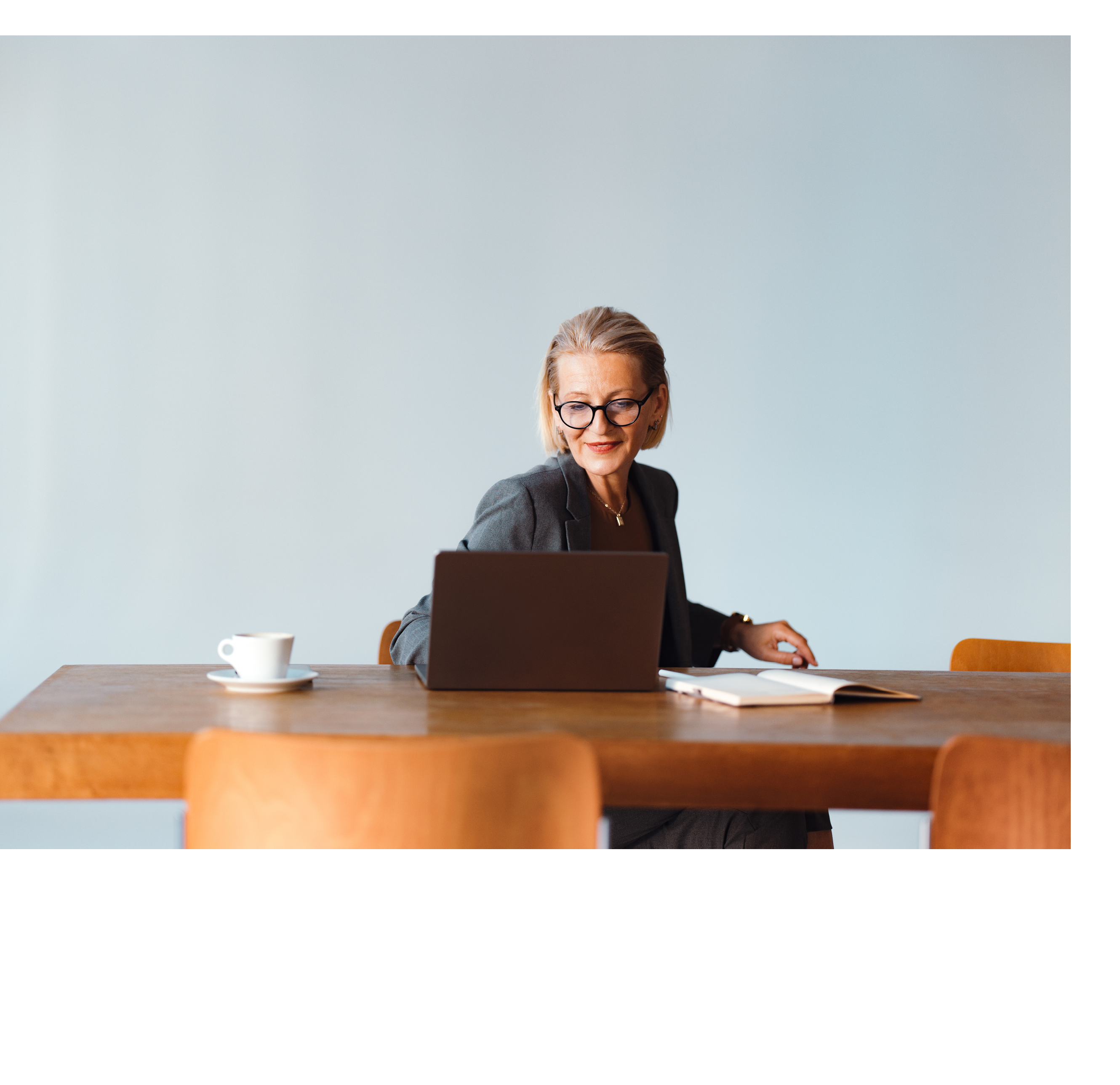 Beautiful senior woman working in the office. She is using a laptop computer and taking notes in the notepad. There is a coffee cup on the table.