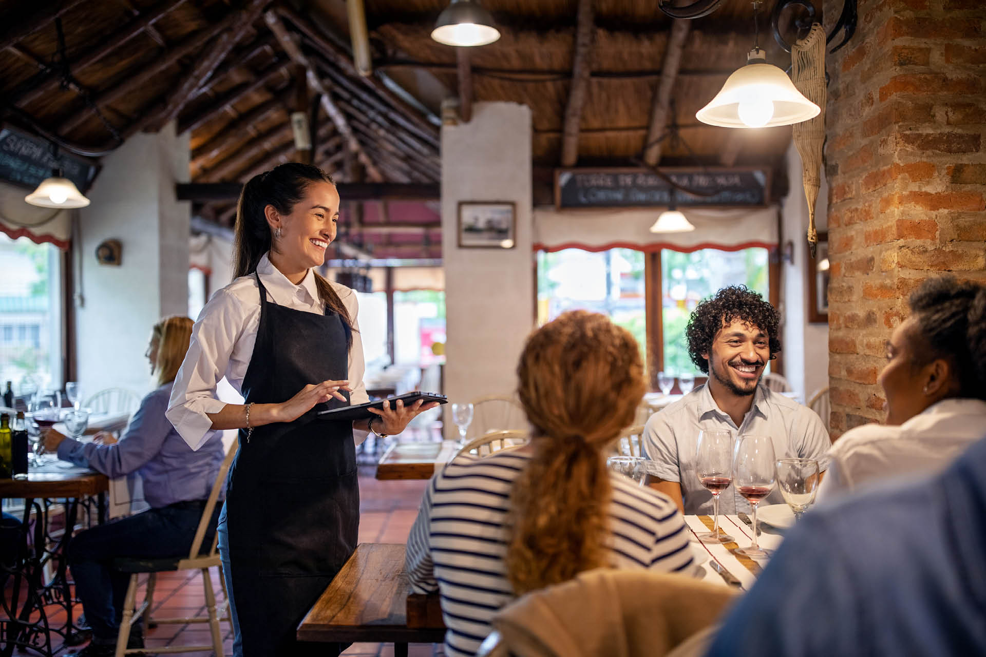 Smiling waitress using a digital tablet while taking lunch order from guests sitting at a restaurant table. Woman working at a restaurant taking order from a group of people.