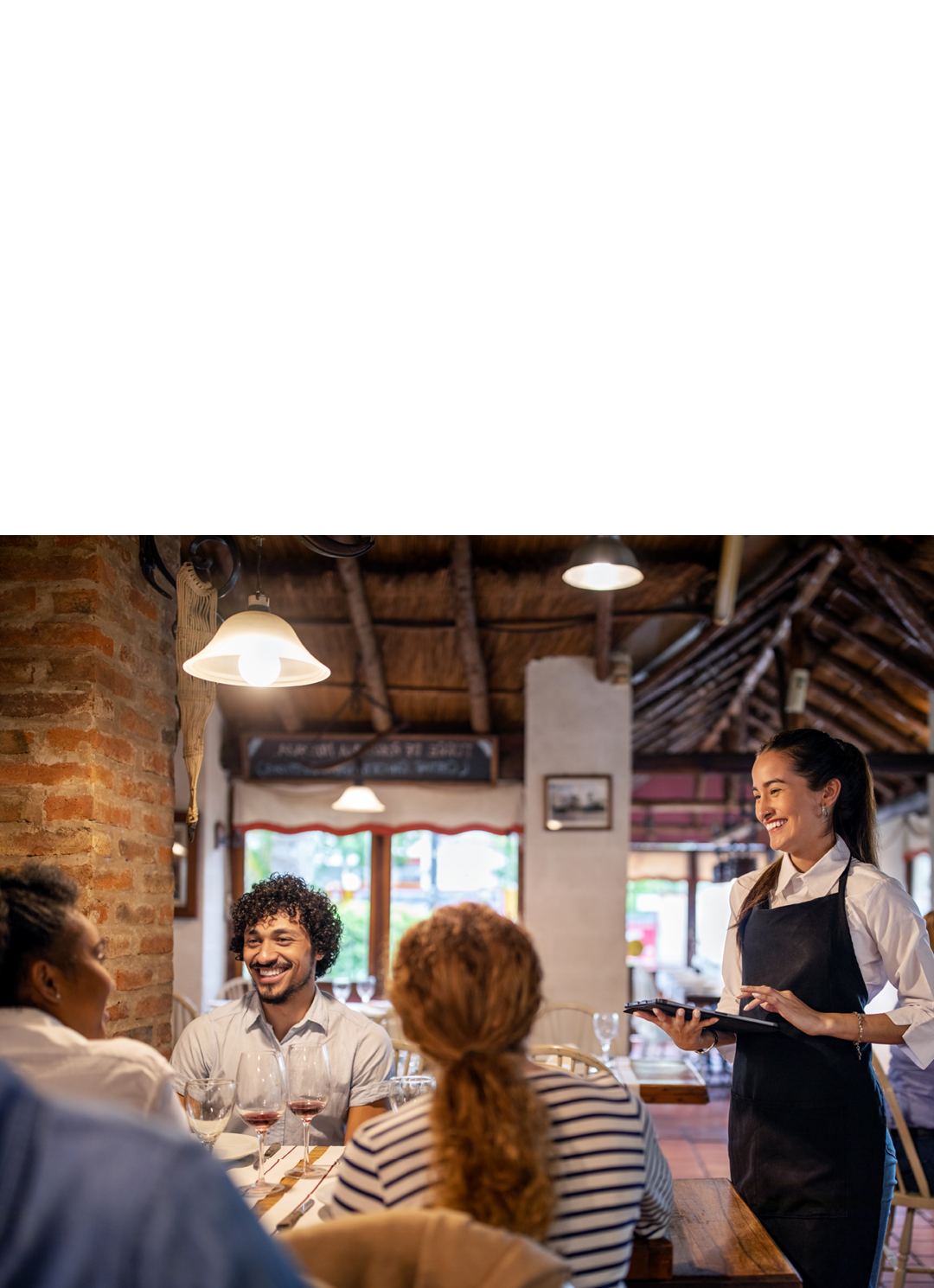 Smiling waitress using a digital tablet while taking lunch order from guests sitting at a restaurant table. Woman working at a restaurant taking order from a group of people.