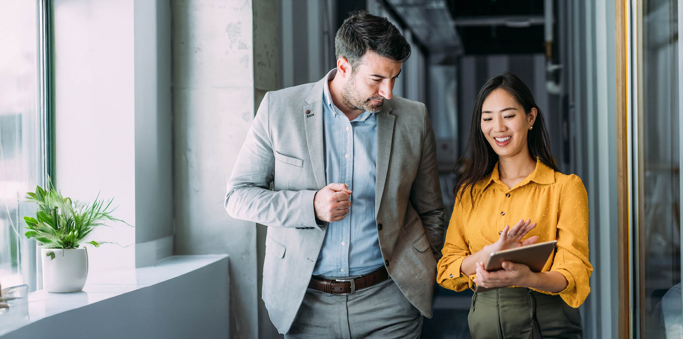 Shot of a two confident business persons talking in the work place. Two colleagues using a digital tablet while walking in a modern office. Businessman and businesswoman in meeting discussing business strategy. Business coworkers working together in the office.