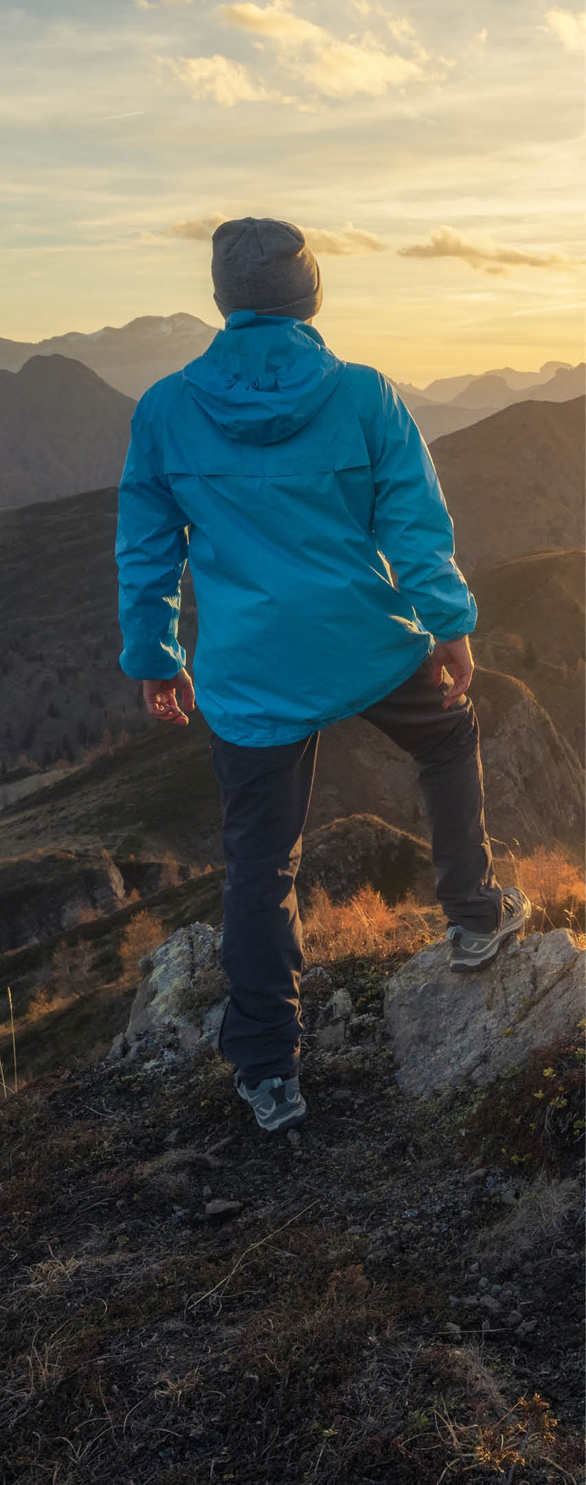 Man on stone on the hill and beautiful mountains in haze at colorful sunset in autumn. Dolomites, Italy. Sporty guy, mountain ridges in fog, orange grass and trees, blue sky with sun in fall. Hiking