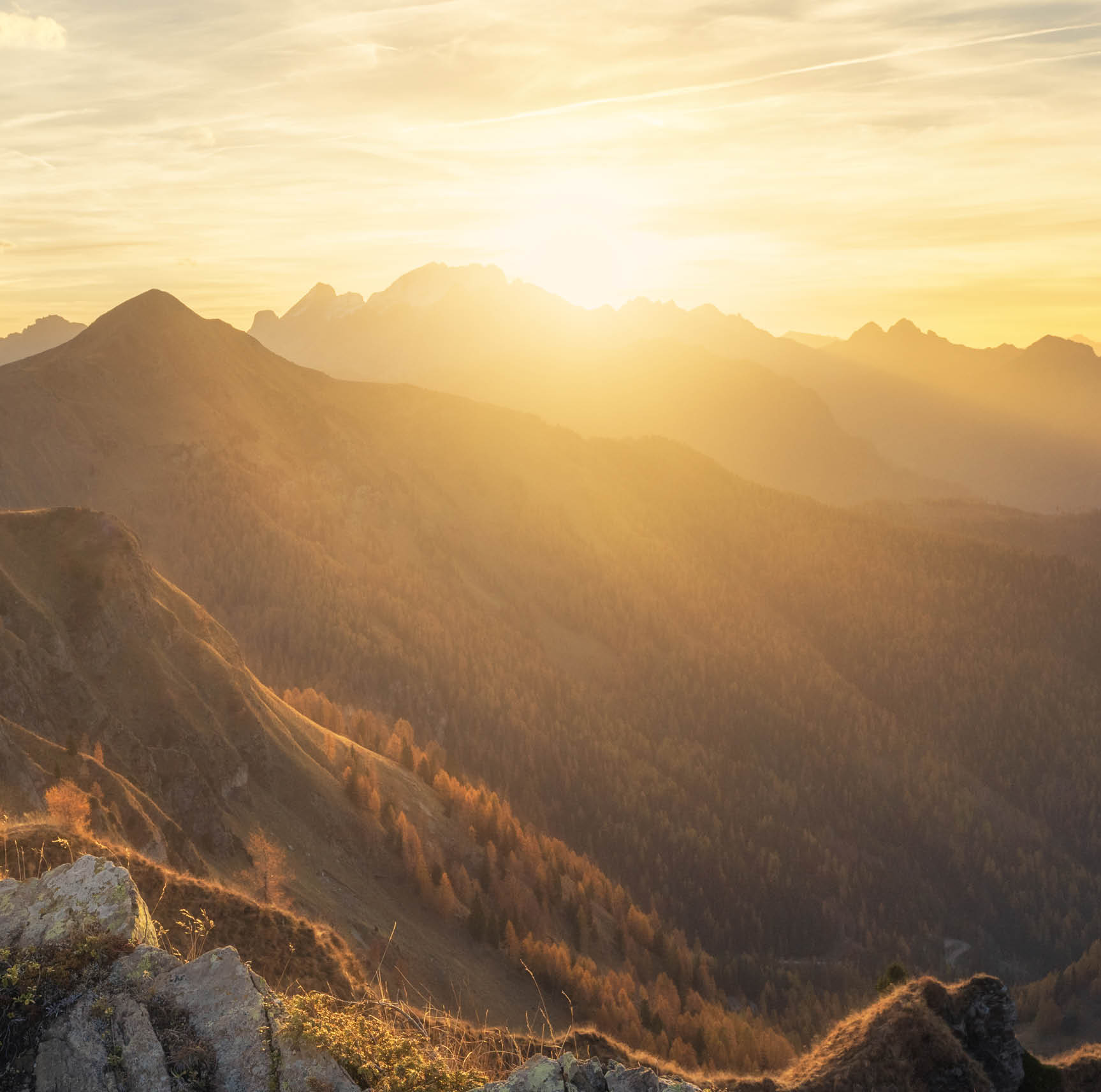 Man on stone on the hill and beautiful mountains in haze at colorful sunset in autumn. Dolomites, Italy. Sporty guy, mountain ridges in fog, orange grass and trees, blue sky with sun in fall. Hiking