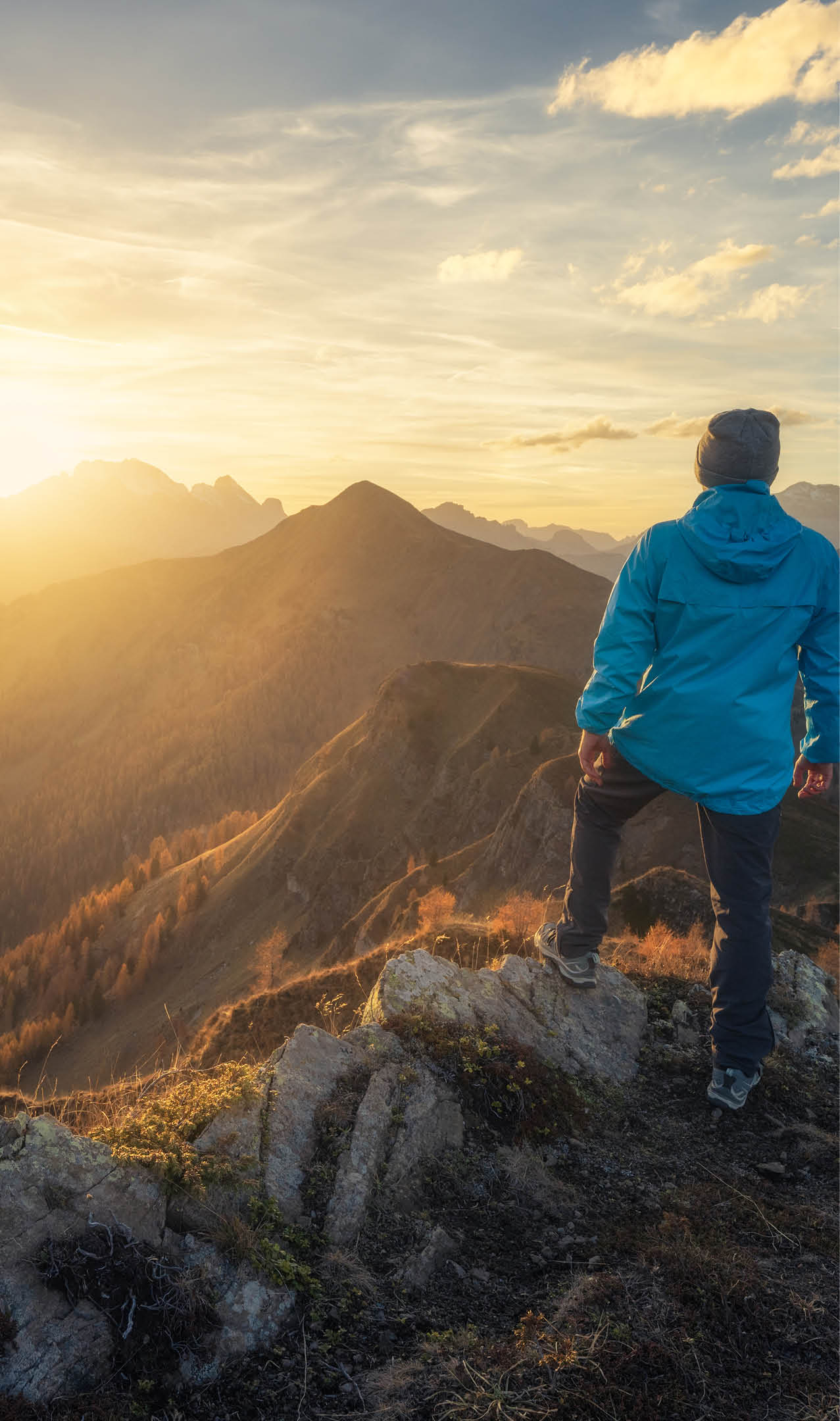 Man on stone on the hill and beautiful mountains in haze at colorful sunset in autumn. Dolomites, Italy. Sporty guy, mountain ridges in fog, orange grass and trees, blue sky with sun in fall. Hiking