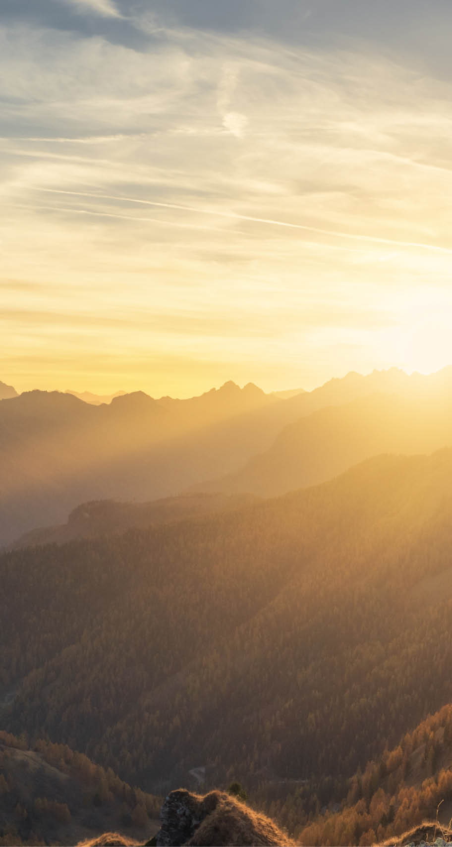 Man on stone on the hill and beautiful mountains in haze at colorful sunset in autumn. Dolomites, Italy. Sporty guy, mountain ridges in fog, orange grass and trees, blue sky with sun in fall. Hiking