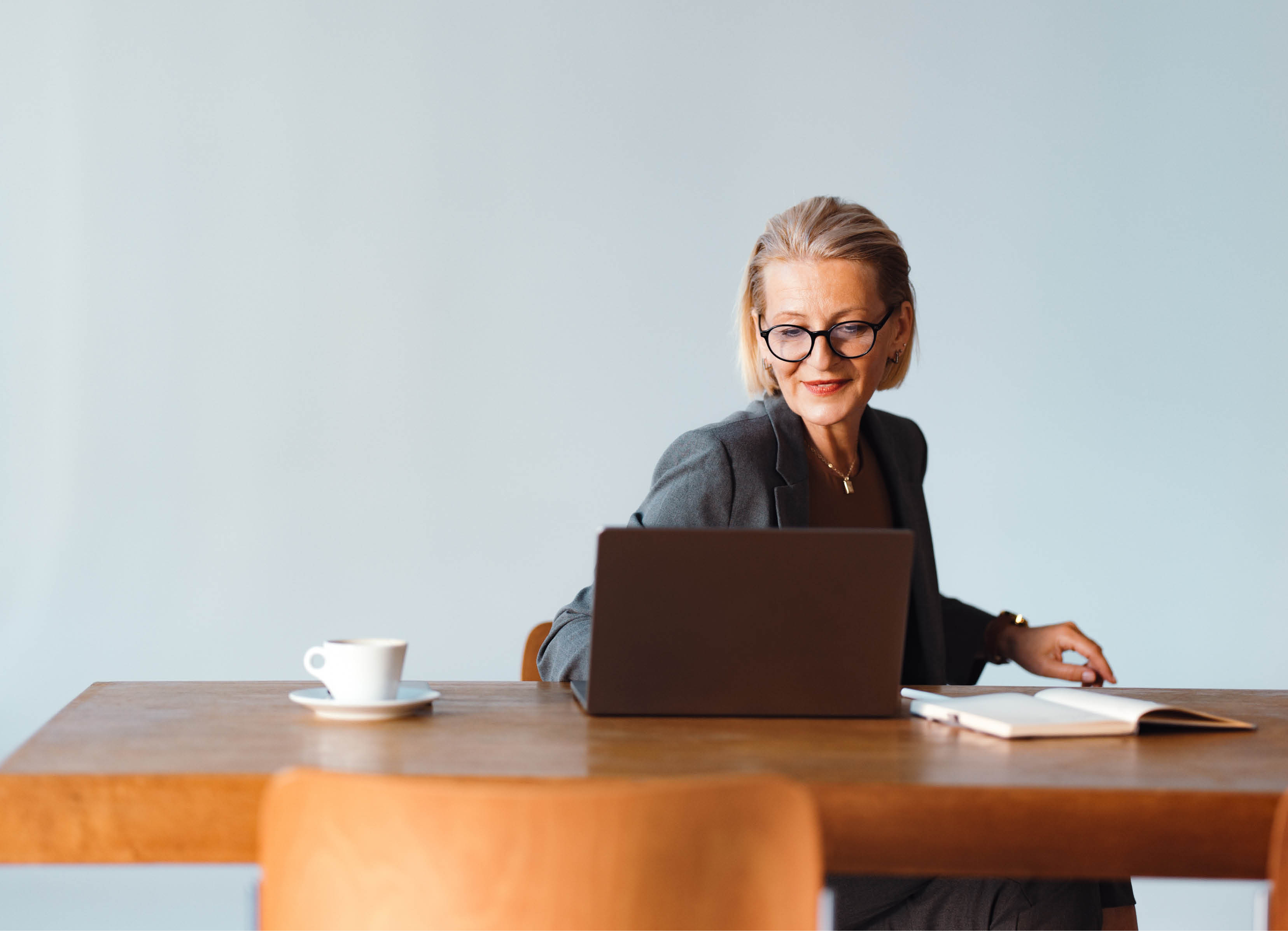Beautiful senior woman working in the office. She is using a laptop computer and taking notes in the notepad. There is a coffee cup on the table.