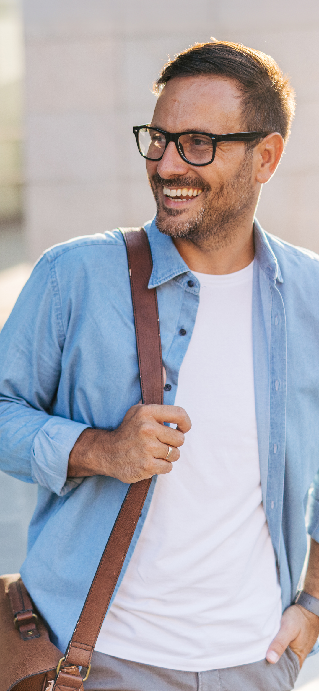 A handsome, cheerful, smiling man walking by a building, carrying bag.