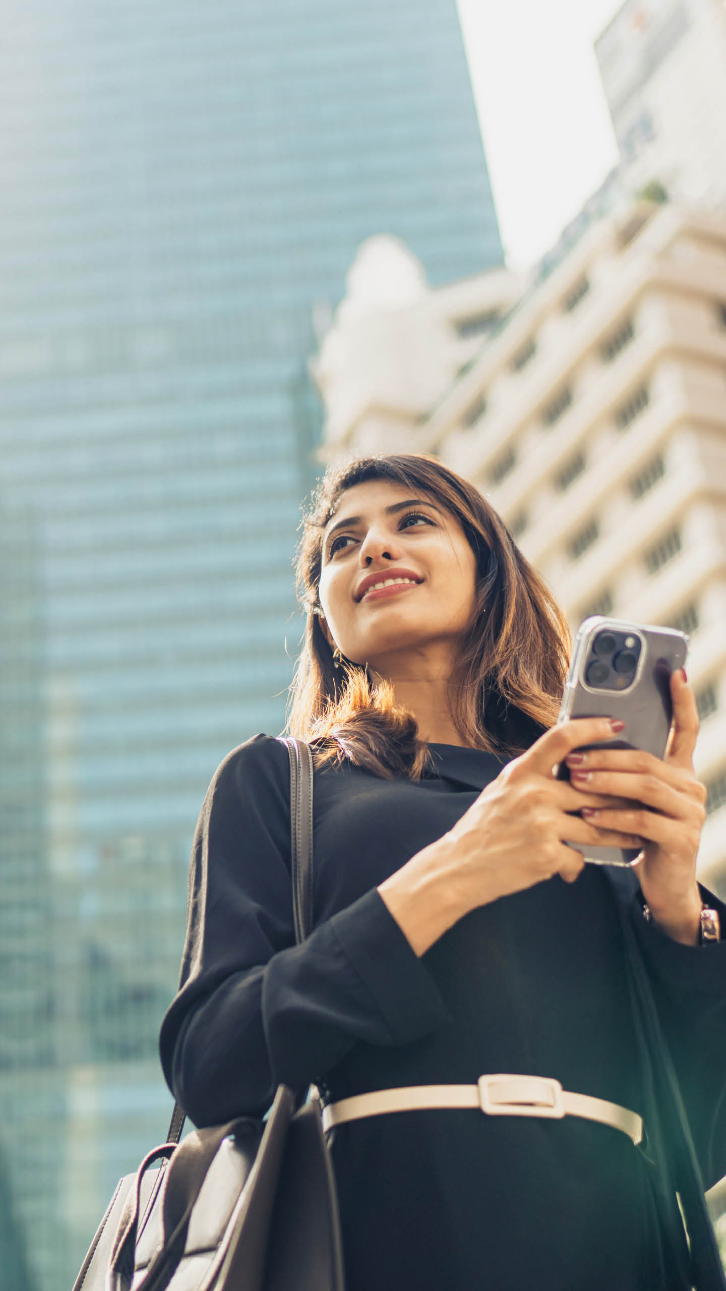 Confident Young Businesswoman With Smartphone Standing Against Modern Buildings