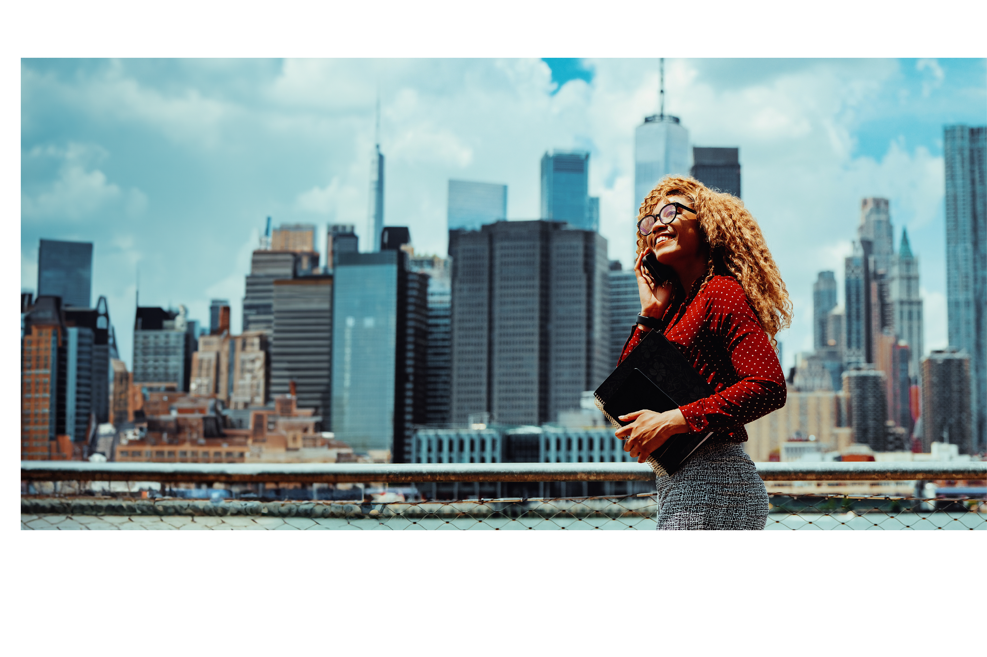 Portrait smiling young adult entrepreneur millennial woman with eyeglasses and afro hair talking on a phone call outdoors with Manhattan New York City skyline skyscraper behind Hudson river